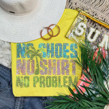 Spangled NO SHOES NO SHIRT NO PROBLEM Transfer, product image showing a vibrant yellow T-shirt with multicolored rhinestone text and palm tree design. Accessories include a wide-brim beige hat, pink glittery hoop earrings, a transparent clutch with gold letters, and a green plant, arranged with denim and striped fabrics for a summery look.