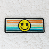 Retro Happy Face Embroidered HAT/POCKET Patch, colorful embroidered patch with a yellow smiley face having star-shaped eyes on a striped rainbow background.