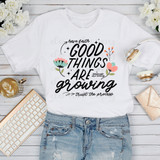 Good Things Are Growing Sublimation Transfer, white T-shirt with floral and motivational text design laid flat on a white wooden surface with a keyboard, cup of coffee, headphones, and a clutch nearby.