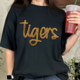 Tiger Gold Sequin Patch, a metallic gold and black embroidered patch with the word 'tigers' in cursive script on a black t-shirt.