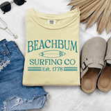Beach Bum Surfing Co DTF Heat Transfer, yellow T-shirt with surfboard graphic and text, surrounded by sunglasses, denim shorts, slippers, and tropical leaves.