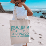 Beach Bum Surfing Co DTF Heat Transfer, beige tote bag with turquoise surf-inspired logo reading 'Beach Bum Surfing Co Est. 1778' held by a woman on a sandy beach