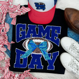 Royal Chenille Football Game Day with Sequin Bow Patch, close-up of a black shirt with blue and white embroidered text and a sequined football bow patch, surrounded by sneakers, a cap, a football, and striped ribbons.