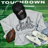 Devils Silver Sequin Patch, a shiny sequin patch featuring the word 'devils' in a stylized script on a light gray sweatshirt surrounded by sneakers, a brown football, a black cap with football embroidery, and casual denim shorts on a green textured background.