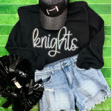 Knights Sequin Patch, a sparkling black patch with silver sequins shaped as a knight's helmet, displayed on a black sweatshirt with the word 'knights' in glittery script, laid out with denim shorts on a green textured surface.