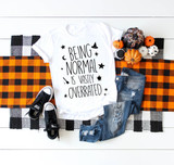 Being normal is vastly overrated Halloween town sublimation transfer, featuring a white t-shirt with Halloween themed text and graphics, paired with denim jeans, black sneakers, and surrounded by autumnal gourds on a black and orange checkered fall-themed blanket.