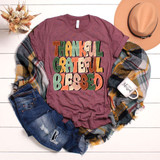 Thankful Grateful Blessed Retro DTF Heat Transfer T-shirt with colorful lettering on a maroon fabric, accompanied by denim shorts, a plaid scarf, a ceramic mug, and a beige hat on a white wooden background.