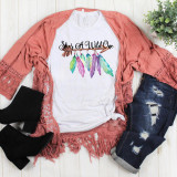 She's a wild one Arrow feather boho sublimation transfer on a white t-shirt with colorful feathers and text, displayed with distressed jeans, black cowboy boots, and a pink lace kimono, on a white wooden background.