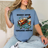 Whiskey Sippin' Turquoise Drippin' Heat Transfer, colorful graphic of a whiskey bottle, glass, and jewelry on a blue t-shirt worn by a smiling woman.