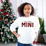 Very Merry Mini Buffalo Plaid DTF Heat Transfer, Christmas-themed sweatshirt with red and black plaid design text, worn by a girl in front of a decorated Christmas tree.