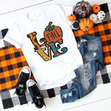Love Fall Sublimation Transfer, festive fall-themed design on a white t-shirt featuring colorful autumn elements and text, surrounded by small decorative pumpkins and plaid patterned cloth with jeans and sneakers.