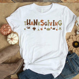 Thanksgiving word art dangle sublimation transfer - Image of a white T-shirt with Thanksgiving themed word art and hanging icons like leaves, pumpkins, and hats, surrounded by fall decorations including mini pumpkins and boots.