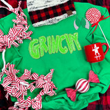 Grinchy Sequin Patch, festive holiday display featuring a green sweatshirt with 'GRINCH' written in sparkly green sequins, surrounded by red and white striped bows, candy-themed decorations, a red mug, a striped hat, and a plush candy ornament, perfect for Christmas fashion or costume accessories.