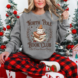 North Pole Book Club DTF Heat Transfer, Christmas-themed sweatshirt displaying a gingerbread man in a cup of hot cocoa, stacked books, holly, and decorative elements, worn by a smiling woman sitting in front of Christmas trees.