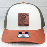 I Do It For The Ho's Leather Hat Patch, leather patch with Santa's face wearing sunglasses and a beard, with the text I Do It For The Ho's, on a trucker hat featuring a white front panel, grey mesh back, and rust-colored brim.