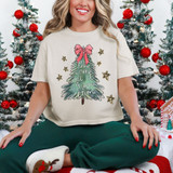 Oh Come Let Us Adore Him Hand Drawn Screen Print Heat Transfer, Christmas themed product featuring a woman sitting in front of decorated Christmas trees, wearing a cream-colored T-shirt with a festive green tree illustration and the phrase 'Oh Come Let Us Adore Him' with a pink bow at the top.