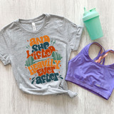 And She Lifted Heavily Ever After DTF Heat Transfer, colorful heat transfer design printed on a gray T-shirt with workout-themed typography and graphics, laid flat on a wooden surface with a purple sports bra and a mint green shaker bottle nearby.