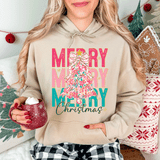 Stacked Merry Christmas DTF Heat Transfer, colorful Christmas tree design on a beige hoodie worn by a woman holding a red mug with whipped cream, with holiday decorations in the background.