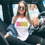 Bright Groovy Mom Faux Sequins DTF Heat Transfer, a woman wearing a white t-shirt with bright pink, orange, and yellow sequined 'MOM' letters, sitting in a car holding an iced coffee, with a beach and palm tree in the background.
