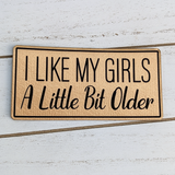 Product: I Like My Girls A Little Bit Older Faux Leather Patch, a beige furry patch with black embroidered text and a black border, placed on a white wooden surface.