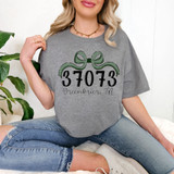 Greenbrier, TN Zip Code Bow DTF Heat Transfer on a gray T-shirt worn by a woman, featuring a green bow design with the zip code 37073 and text 'Greenbrier, TN' underneath.