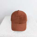 Brown Corduroy Hat, a close-up image of a stylish brown corduroy baseball cap resting on a white textured surface against a light-colored wall background.