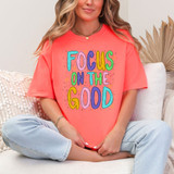 Focus On The Good Stars DTF Heat Transfer, colorful motivational quote on a coral t-shirt worn by a woman sitting cross-legged on a sofa.