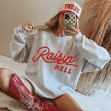 Raisin' Hell DTF Heat Transfer, a close-up of a woman wearing a gray sweatshirt with the red text 'Raisin' Hell' and a white and red cap, taking a mirror selfie with a checkered phone case.