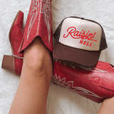 Raisin' Hell DTF Heat Transfer, red cowboy boots, and a cap with Raisin' Hell logo on a white fuzzy surface.