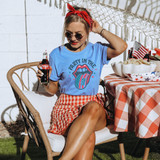 Party In The USA Tounge DTF Heat Transfer, woman wearing a blue T-shirt with patriotic design, red bandana, sunglasses, sitting outdoors at a patriotic party with American flags and red, white, and blue decorations.
