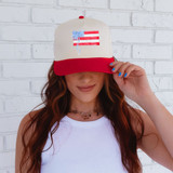 Coquette USA Flag DTF Heat Transfer, product image of a woman wearing a trucker hat with an American flag design and a blue bow, against a white brick wall background.