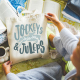 Jockeys, Derby Day & Juleps DTF Heat Transfer, printed on a tote bag, surrounded by magazines, a cup of coffee, and clothing on a bed.