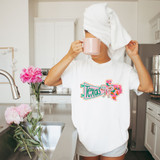 Texas State Pride Bow DTF Heat Transfer, vibrant and colorful design featuring the shape of Texas with floral patterns and a bow, applied on a white t-shirt worn by a woman with a towel wrapped on her head, holding a pink mug in a kitchen setting.