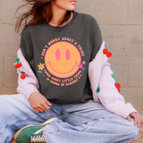 Don't Worry About A Thing Retro Happy Face DTF Heat Transfer, a woman wearing a dark grey sweatshirt with a pink smiling face graphic and a motivational quote, paired with light jeans and green sneakers, sitting against a beige brick wall.