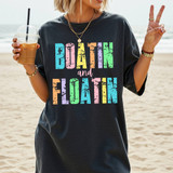 Distressed Boatin And Floatin DTF Heat Transfer, colorful distressed text design on a dark T-shirt showing a woman at the beach with a drink making a peace sign gesture.