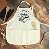 Watch Me Whip It FINAL STOCK Screen Print Transfer, an apron with a whip illustration and text design, on a wooden surface with cookies and kitchen utensils around it.