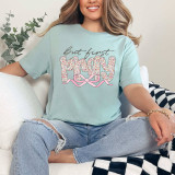 But First Pray Coquette DTF Heat Transfer, woman wearing a light blue t-shirt with floral 'Mommy' design and black script text, sitting on a white couch in a cozy room.