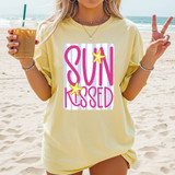 Striped Sunkissed DTF Heat Transfer, beach scene with a woman wearing a yellow T-shirt featuring pink and lavender striped design with starfish accents and the words 'Sun Kissed' in bold pink letters.