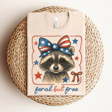 Feral But Free Raccoon DTF Heat Transfer, a cute raccoon illustration with a patriotic red, white, and blue bow and stars, on a beige T-shirt.
