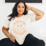 He Has A Plan I Have A Purpose Happy Face ORANGE DTF Heat Transfer, woman wearing a beige T-shirt with orange smiley face, star, and lightning bolt design and text, sitting on a bed.