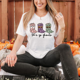 Let's Go Ghouls Boots DTF Heat Transfer, woman wearing a Halloween-themed T-shirt with colorful ghoulish boots illustration and the text 'let's go ghouls', sitting among pumpkins.