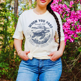 When You Pass Through Deep Waters DTF Heat Transfer, image of a woman wearing a cream T-shirt with a turtle and ocean design and motivational quote, outdoors with pink flowers and greenery.