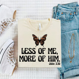 Less Of Me, More Of Him Butterfly DTF Heat Transfer, white T-shirt with butterfly graphic and biblical quote, casual jeans, sneakers, and jewelry on a white quilted surface.