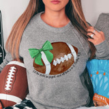 Let The Field Be Joyful Football DTF Heat Transfer, image of a woman wearing a gray sweatshirt with a football graphic and a green bow, holding a football and sitting beside a blue snack bag.