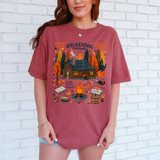 Cozy Cabin Reading Retreat DTF Heat Transfer, a graphic t-shirt featuring a cozy cabin surrounded by trees, pumpkins, a campfire, books, and autumn-themed decorations, perfect for fall reading and relaxation.