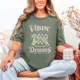 Vibin' With My Demons DTF Heat Transfer, a Halloween-themed graphic design T-shirt featuring green zombies' hands, skeleton finger, bats, and stars on an olive green fabric.
