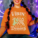 Vibin' With My Demons DTF Heat Transfer product, orange t-shirt with Halloween-themed graphic and text, zombie hand and skeleton hand, stars, and bats, worn by a smiling woman with braided hair, halloween party background.