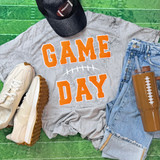 Game Day with Silicon Laces Patch - 10 Colors, sports-themed casual outfit with a gray T-shirt displaying 'Game Day' in orange lettering, a black cap, white sneakers, blue jeans, and a brown tumbler with a football-themed design.
