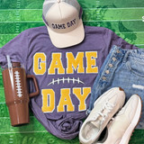 Game Day with Silicon Laces Patch - 10 Colors, sports-themed apparel and accessories including a purple T-shirt with 'GAME DAY' in yellow lettering and a fishbone graphic, a white cap with 'GAME DAY' patch, white sneakers, a brown tumbler with white stitch pattern, and denim shorts, all arranged on a green textured background.