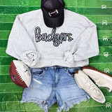 Badgers Sequin Patch, embroidered patch on grey sweatshirt with football helmet, sneakers, and denim shorts on a green football field background.
